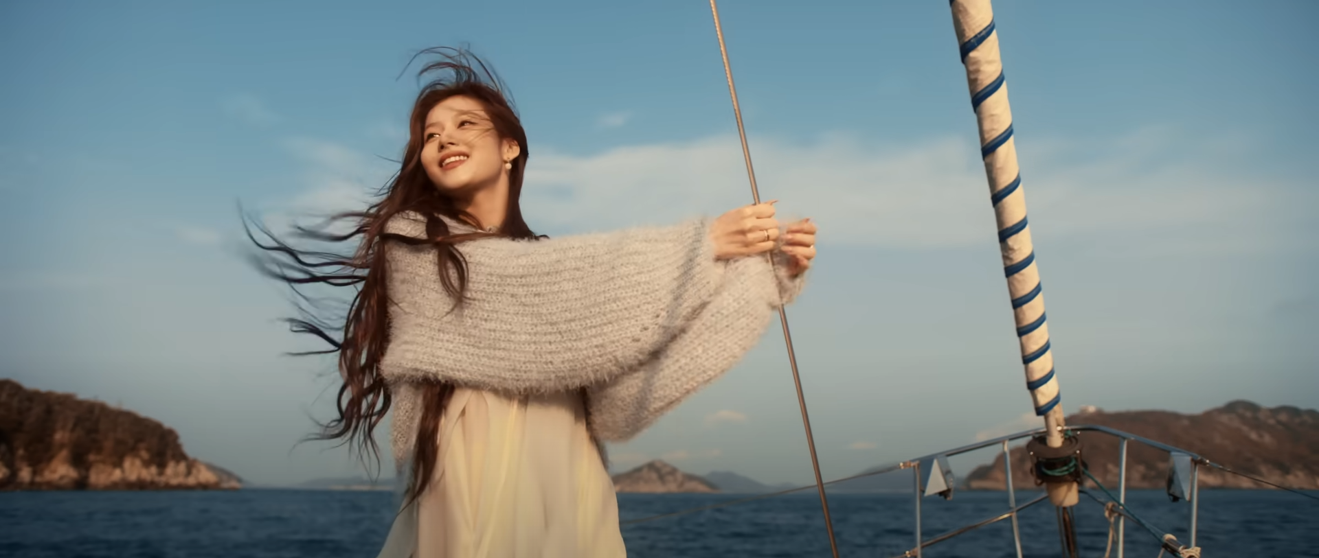 a woman with long brown hair stands on a sailboat, her arms outstretched as if embracing the wind. she's wearing a light blue chunky knit sweater over a flowing white dress. the sea stretches out before her, dotted with islands bathed in the warm golden light of the setting sun. the sky is a vibrant blue, with fluffy white clouds scattered across it. the overall feeling of the image is one of joy, freedom, and connection with nature.