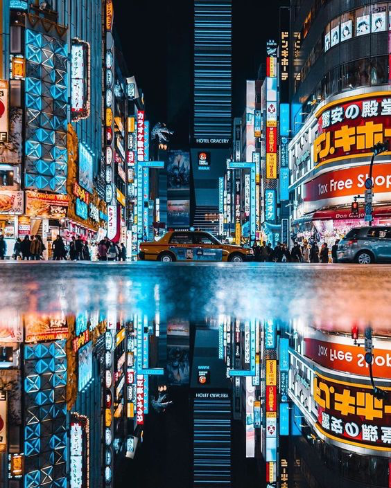 the image is a photograph of a bustling street in tokyo, japan, at night. the street is lined with tall buildings with brightly lit neon signs. the image is symmetrical, with the top half of the image showing the street and the bottom half showing its reflection in a puddle of water on the ground. a taxi is driving down the street in the center of the image. people are walking on the sidewalks on either side of the street. the overall atmosphere is one of energy and excitement.