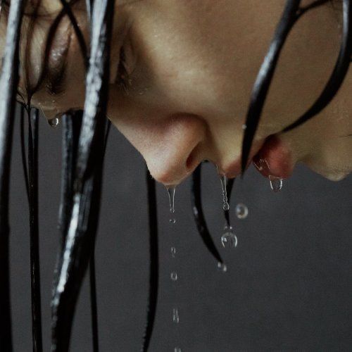 the image is a closeup portrait of a woman's face with long, dark, wet hair.  water is dripping from her hair and chin, suggesting she has just washed or been in the rain. her expression is serious, almost melancholic, with her eyes downcast. the overall mood is dark and contemplative, with a focus on texture and contrast. the dark background accentuates the water droplets and the woman's features. the photograph has a stylistic and artistic quality, with a subdued color palette.