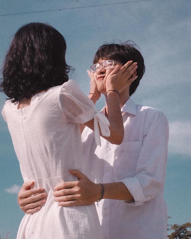 a young couple is hugging each other while looking into each other's eyes. the girl is wearing a white dress and the boy is wearing a white shirt. the background is a blue sky.  the image portrays a romantic and affectionate moment between the couple.