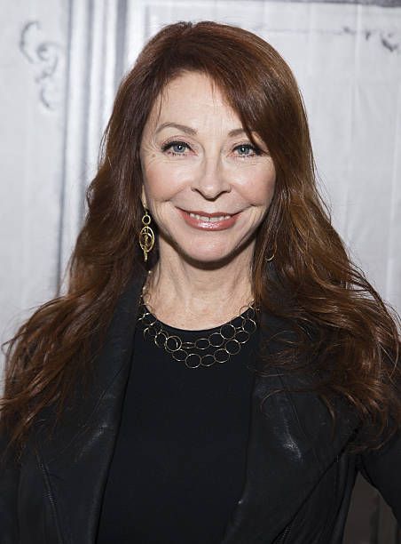 a woman with long, dark hair smiles at the camera. she is wearing a black leather jacket, a black shirt, a gold necklace, and gold earrings. the background is a white wall with a slight texture.