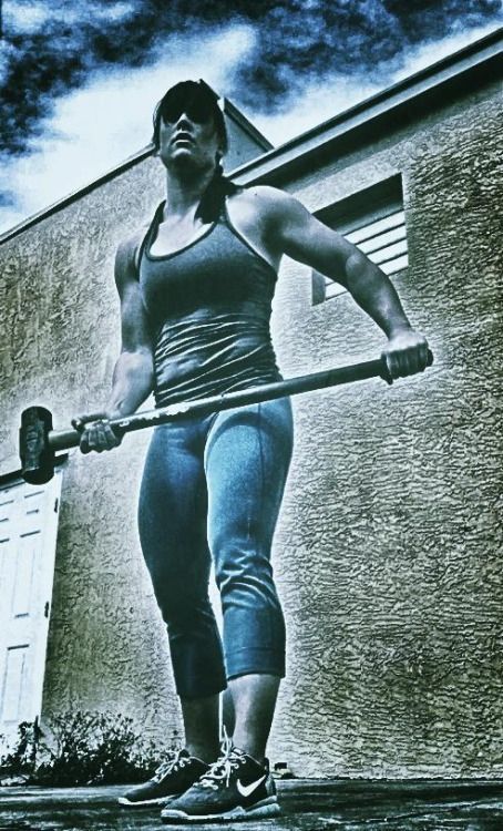 a woman is holding a sledgehammer in a gym setting. she is looking determined and ready to work out.  she is wearing athletic clothing and shoes, ready to train.  the image is a little grainy, like a photo taken on a smartphone.  the colors are muted, with a blueish cast.