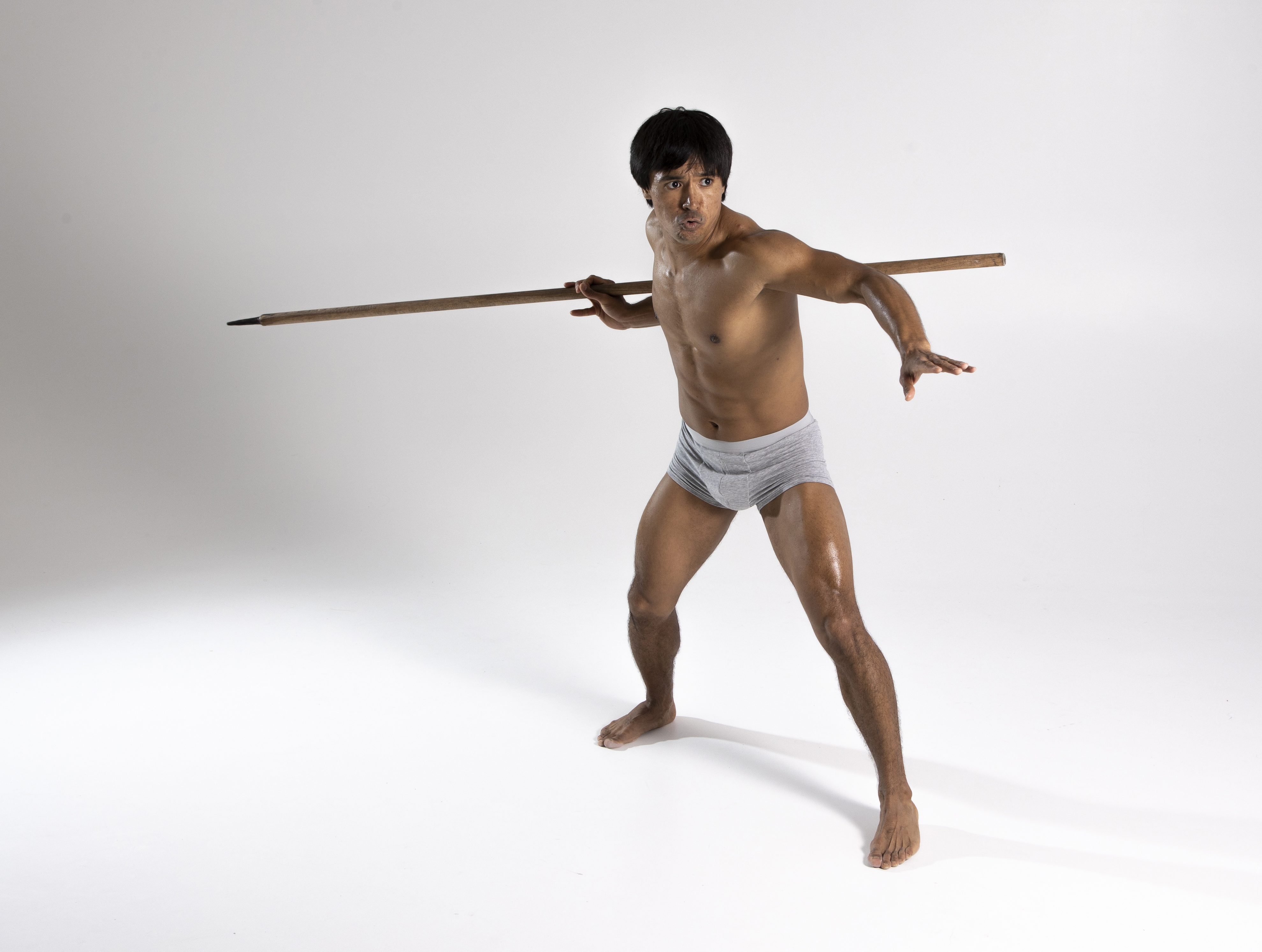 a shirtless man in grey underwear stands against a white background, holding a wooden spear horizontally across his shoulders. he strikes an action pose, with an intense expression on his face. his physique is athletic and he is barefoot. the shot is a full shot taken in a studio.