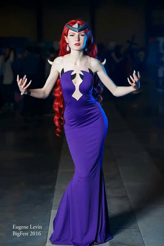 a woman in a cosplay costume stands in a convention center. she wears a long, formfitting purple dress with black accents and a cutout at the chest. she has red hair styled in curls, and she wears a black headpiece with horns. she also has dangling earrings and long, white nails. the background is a dimly lit convention center with other people in the distance.