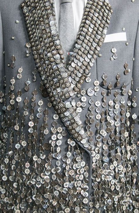 a closeup shot of a grey suit jacket adorned with a pattern of silver buttons. the buttons are sewn onto the fabric in a cascading pattern, creating a unique and interesting texture. the jacket is welltailored and has a classic style. the buttons are a statement piece and add a touch of glamor to the outfit. the image is a beautiful example of how simple materials can be used to create something striking.
