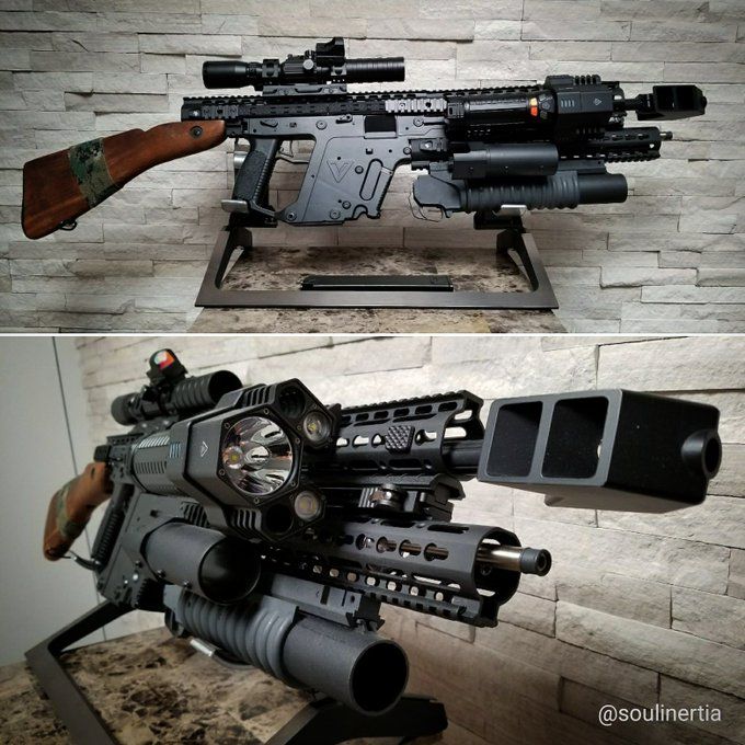two images of a highly modified rifle, likely an ar15 variant. the rifle is black with a wooden stock and features numerous attachments including a scope, flashlight, grenade launcher, foregrip, and a large muzzle device. it is displayed on a stand against a wall with a stone pattern. the bottom image provides a closer view of the rifle's front end.