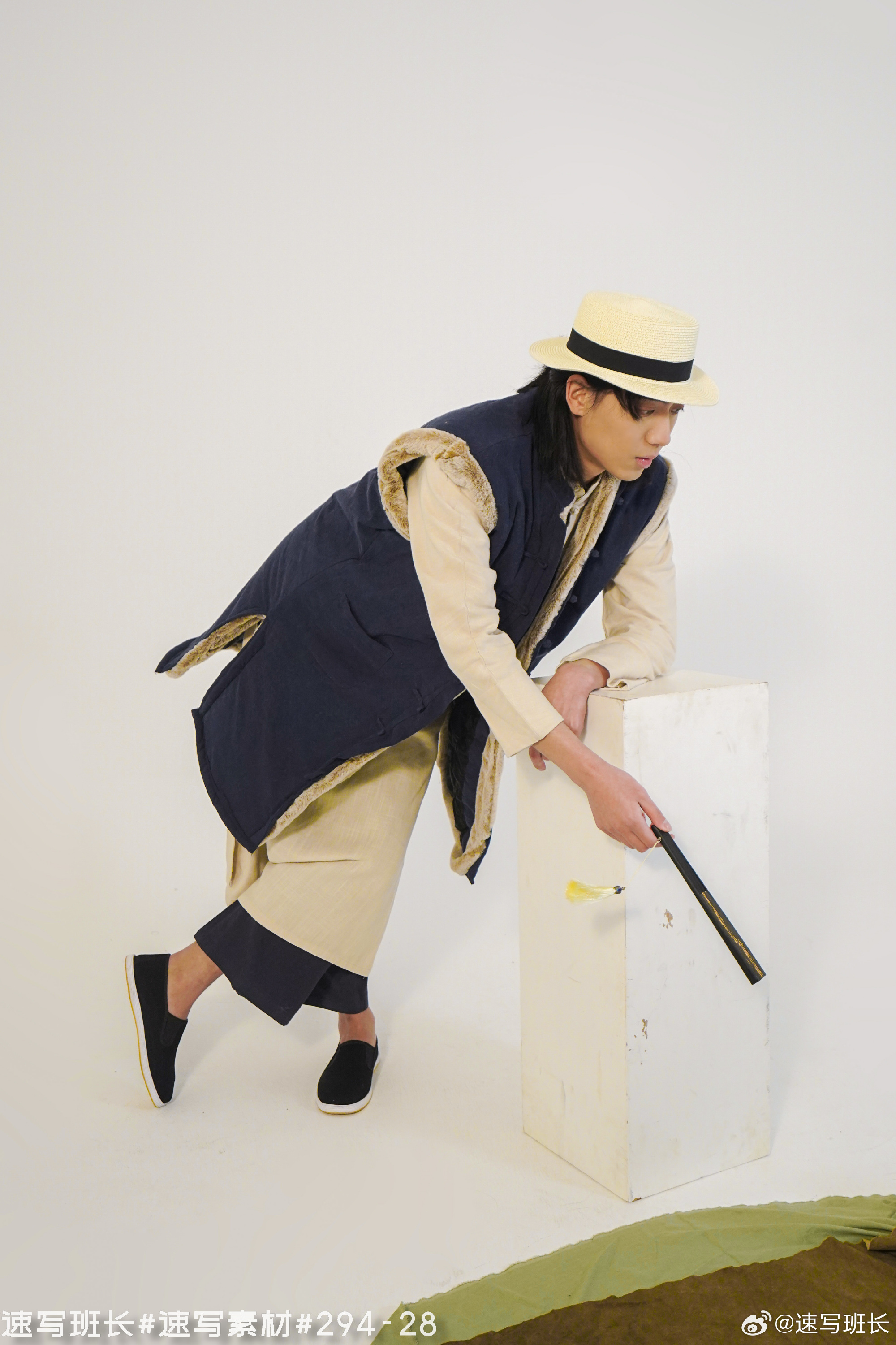 a man is standing, slightly bent over and holding a long, thin black object in his hand. he is wearing a blue vest with a tan fur trim over a long tan shirt and black pants. he is also wearing black shoes and a straw hat. he is posing in a studio setting with a white block in front of him and a green rug on the floor. the lighting is soft and the background is white.
