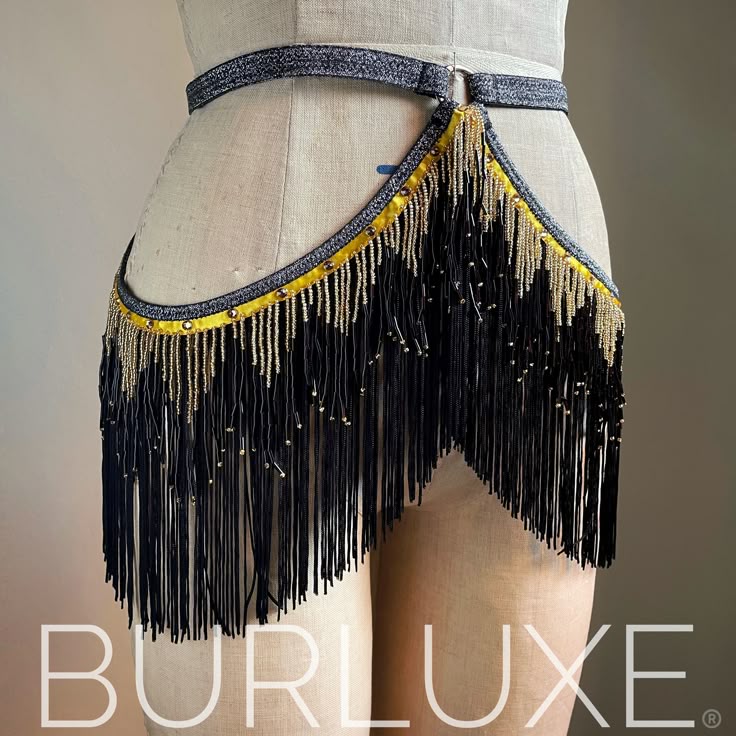 a closeup studio shot of a burlesque belt displayed on a mannequin. the belt features a silver and black glitter belt, a yellow trim with gold studs, and black beaded fringe. the fringe is embellished with gold beads. the brand name "burluxe" is displayed at the bottom of the image.