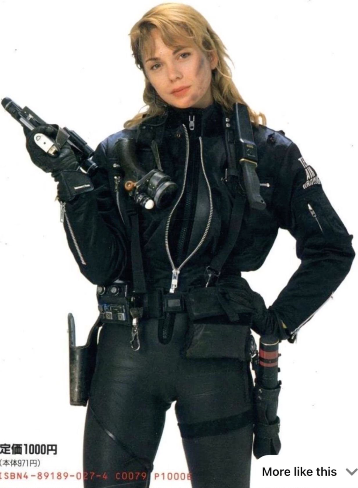 a woman in a black leather jacket holds a gun. she is wearing a lot of tactical gear including a knife on her belt. she is standing in a white studio.