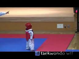 a young boy in a white taekwondo uniform with red headgear and  red chest protector is standing on a blue and red mat. he's looking towards the camera and waiting for instructions from the referee. he's in a taekwondo class,  practicing his martial arts skills.