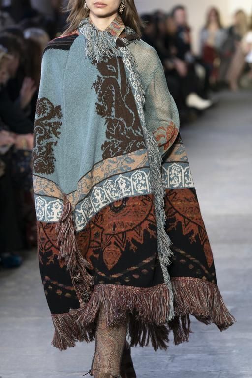a woman in a blue, black and brown patterned poncho with a fringe trim walks down a runway.