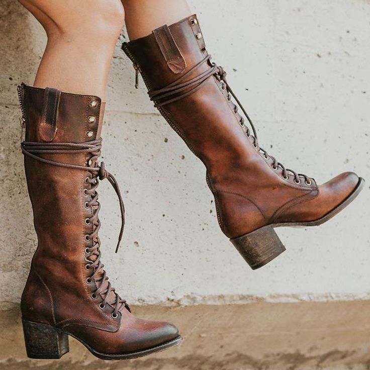 the image shows a pair of women's brown leather kneehigh boots.  the boots are laceup style with a chunky heel and a side zipper.  the leather has a distressed or vintage look, giving them a rustic and worn appearance.  the boots are being modeled by a woman who is sitting with her legs slightly apart, showcasing the boots from multiple angles. the background is a lightcolored, textured wall. the overall style of the boots suggests a bohemian or vintage aesthetic.
