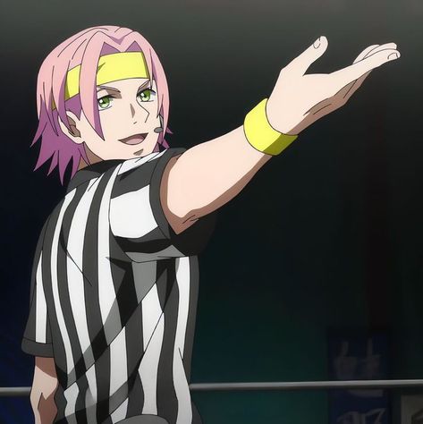 an animestyle image of a young man with short pink hair and a yellow headband acting as a referee. he's wearing a black and white striped referee shirt and has a microphone headset around his ear. he's extending his right arm out in a gesture of announcement or direction, his hand open wide. the background suggests an indoor setting, possibly a boxing ring or wrestling arena, with blurred elements indicating a crowd or equipment.