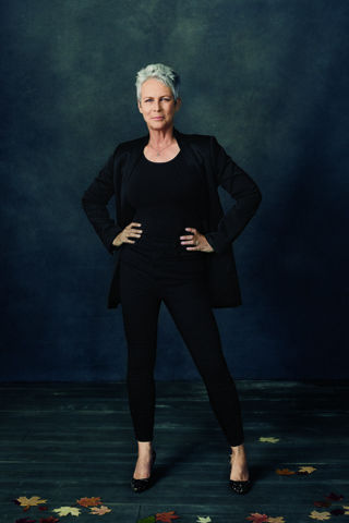 the image is a professional studio portrait of jamie lee curtis. she's standing confidently with her hands on her hips, wearing a black blazer, black top, and black skinny pants. her short gray hair is styled neatly, and she's wearing black high heels. the background is a dark, textured bluegray, with some scattered autumn leaves on the dark wooden floor in the foreground. the overall mood is serious and sophisticated.