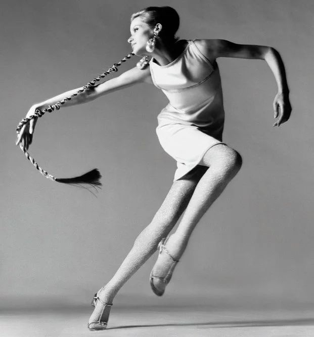 a black and white fashion photograph features a woman in midmotion. she is wearing a simple, sleeveless dress and patterned tights, paired with strapped heels. large, round earrings adorn her ears, and her hair is styled up with a long braid extending from the side of her head. the braid is decorated with what looks like beads, adding to the visual interest. the model is positioned against a plain studio backdrop, which highlights her pose and attire.