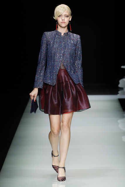a blonde woman in a blue jacket, burgundy skirt, and striped heels poses on a runway, with a black background.