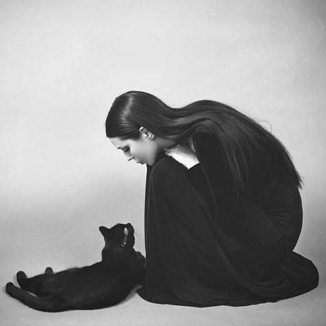 a black and white portrait of a woman with long black hair sitting on the floor and looking down at a black cat. the woman is wearing a black dress, and the cat is lying on its side. the background is a plain white wall.