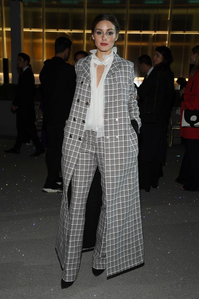 olivia palermo is captured in a full shot, standing confidently.  she's wearing a stylish, long black and white plaid overcoat paired with matching wideleg pants and a white blouse underneath. the outfit is sophisticated and chic. the background shows a blurredout scene suggestive of an evening event or red carpet, with other people in the background.  the overall impression is of high fashion and elegance.