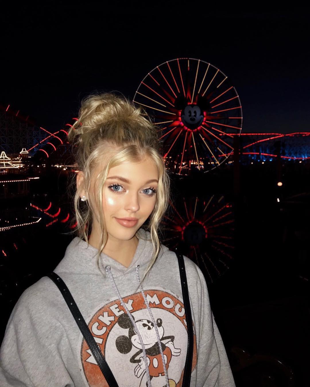 a young woman with blonde hair styled in an updo is posing for a photo in front of a ferris wheel at night. she is wearing a gray hoodie with a mickey mouse design and has on makeup. the ferris wheel is lit up with red lights and has a mickey mouse face in the center. the background is dark with other amusement park rides visible. the woman is also wearing hoop earrings and a necklace.