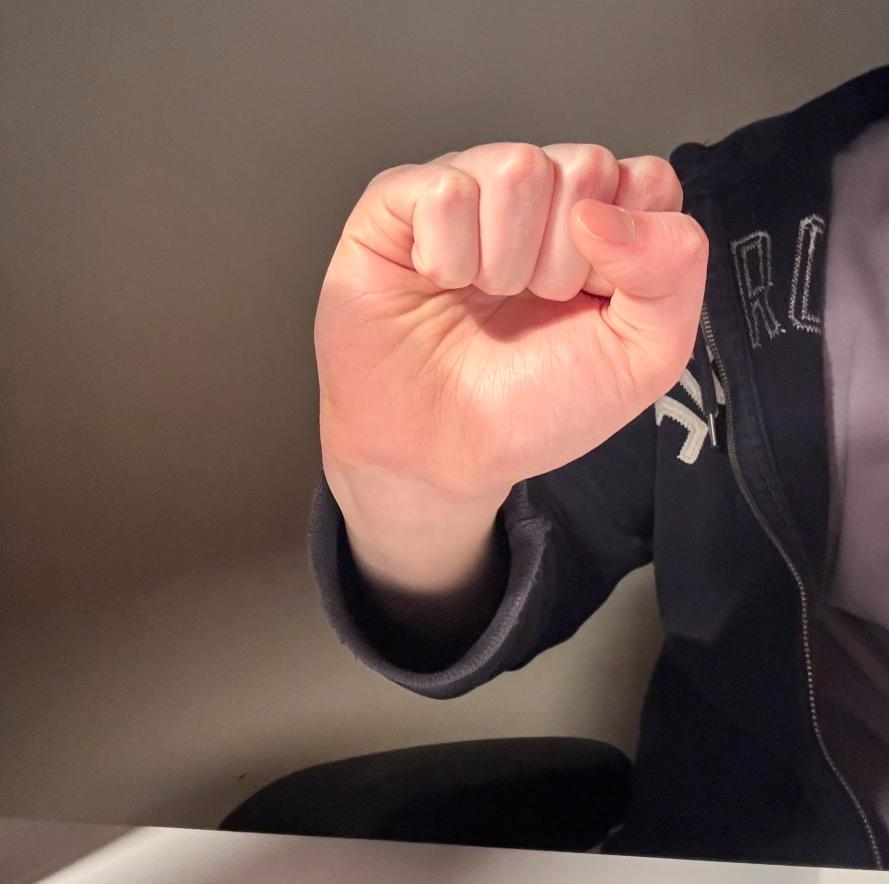 a person's hand is making a fist in front of a white wall. the hand is lit by natural light, casting a shadow on the wall. the person is wearing a black hoodie with white lettering.