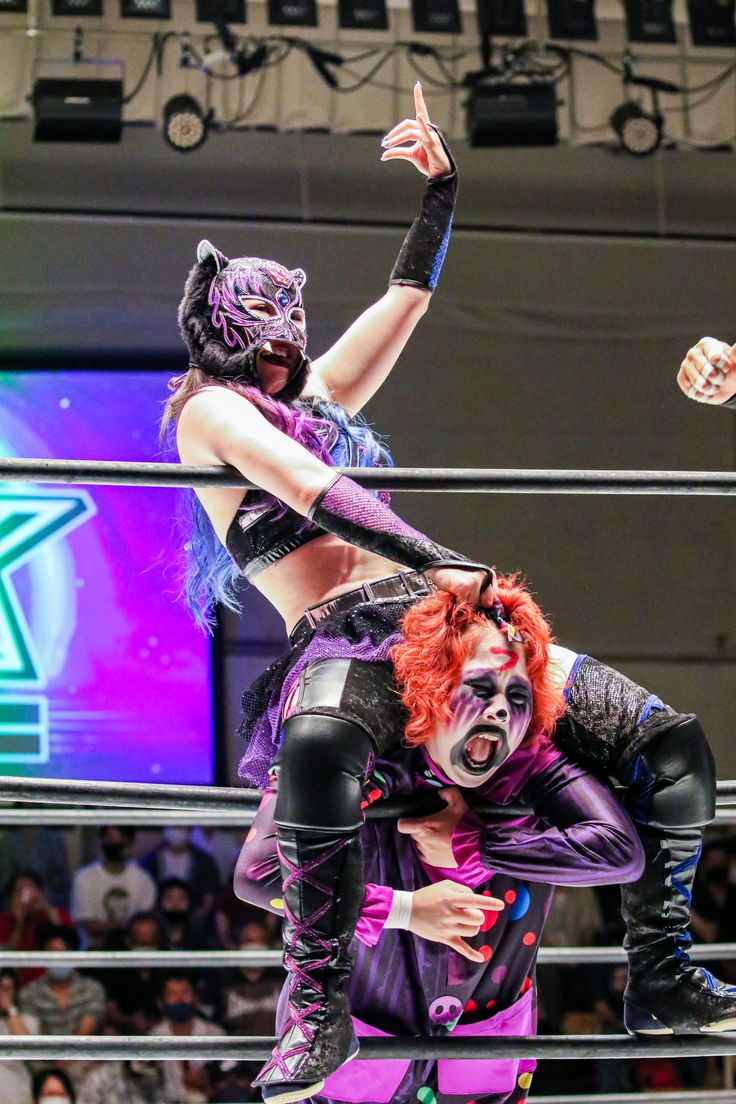 two women wrestlers are in the ring. the woman on top is wearing a purple and black costume with a cat mask. the woman on the bottom is wearing a purple and white costume with a clownlike makeup. they are both in a fighting stance, with the top woman in a position to strike. the audience is in the background watching the match.
