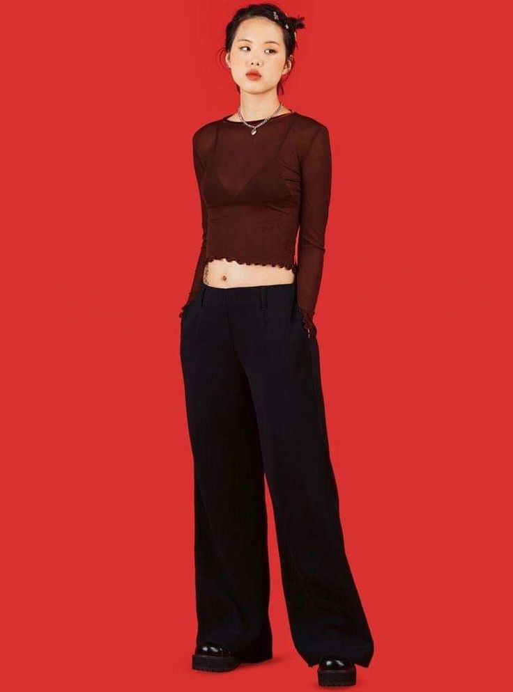 the image shows a young woman standing against a solid red background. she is wearing a brown sheer, longsleeved cropped top over a dark bra, highwaisted wideleg black pants, and black platform shoes. she has a simple choker necklace on. her hair is pulled back. the overall style is a blend of minimalist, dark academia and grunge aesthetics with a hint of y2k influence. the red background provides a stark contrast to her outfit, making the clothing stand out prominently. the pose is casual yet stylish, showcasing the clothing well.
