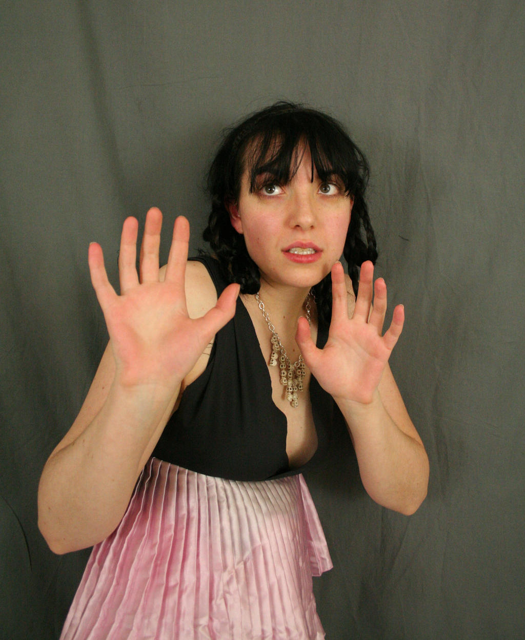 loseup view of a young woman with dark hair and bangs, wearing a black top and pink pleated skirt. she has a look of fear or anxiety on her face, with her hands raised in a defensive or protective gesture. the background is a simple, dark gray backdrop. the image suggests a feeling of vulnerability or apprehension.