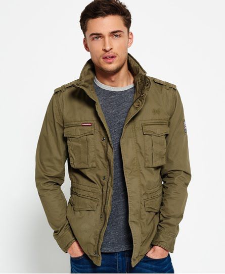 a young man wearing a green military jacket, standing outdoors. he is looking at the camera. the jacket is unbuttoned, and he is wearing a grey tshirt underneath. he is also wearing dark wash jeans.