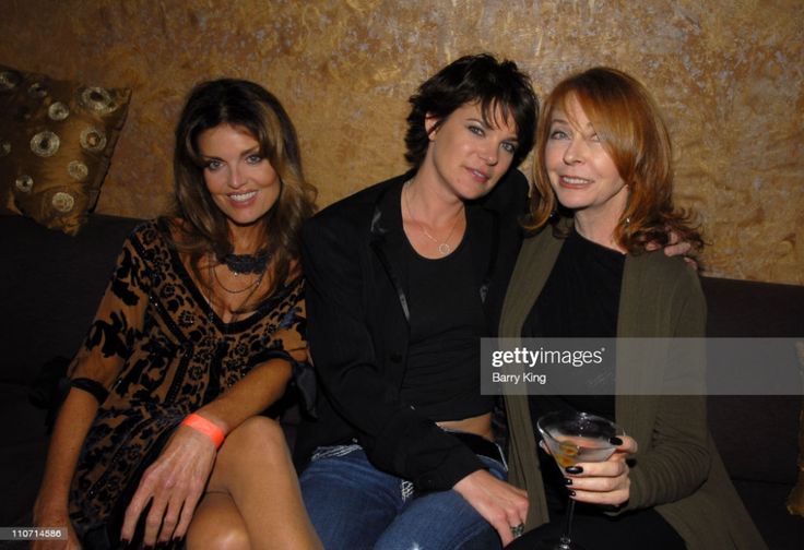 three women pose together on a couch. two are smiling and one is holding a glass of wine. the woman on the left is wearing a black patterned dress and has long, brown hair. the woman in the middle is wearing a black jacket over a black tank top and has short, dark hair. the woman on the right is wearing a green sweater and has long, blonde hair. the woman on the right is smiling while looking at the camera. the other two women are looking at each other and smiling. they are all dressed in fashionable clothes. the lighting is dim. they are all looking happy and are enjoying their time together.