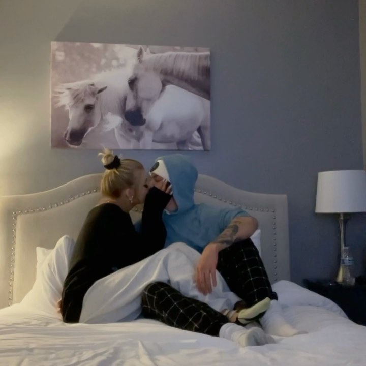 a couple is sitting on a bed in a bedroom, sharing a kiss. the woman has blonde hair in a bun and is wearing a black shirt and plaid pants. the man is wearing a blue hoodie, plaid pants, and socks. he has tattoos on his arm. above the bed is a painting of two white horses. a lamp sits on a nightstand to the right of the bed.