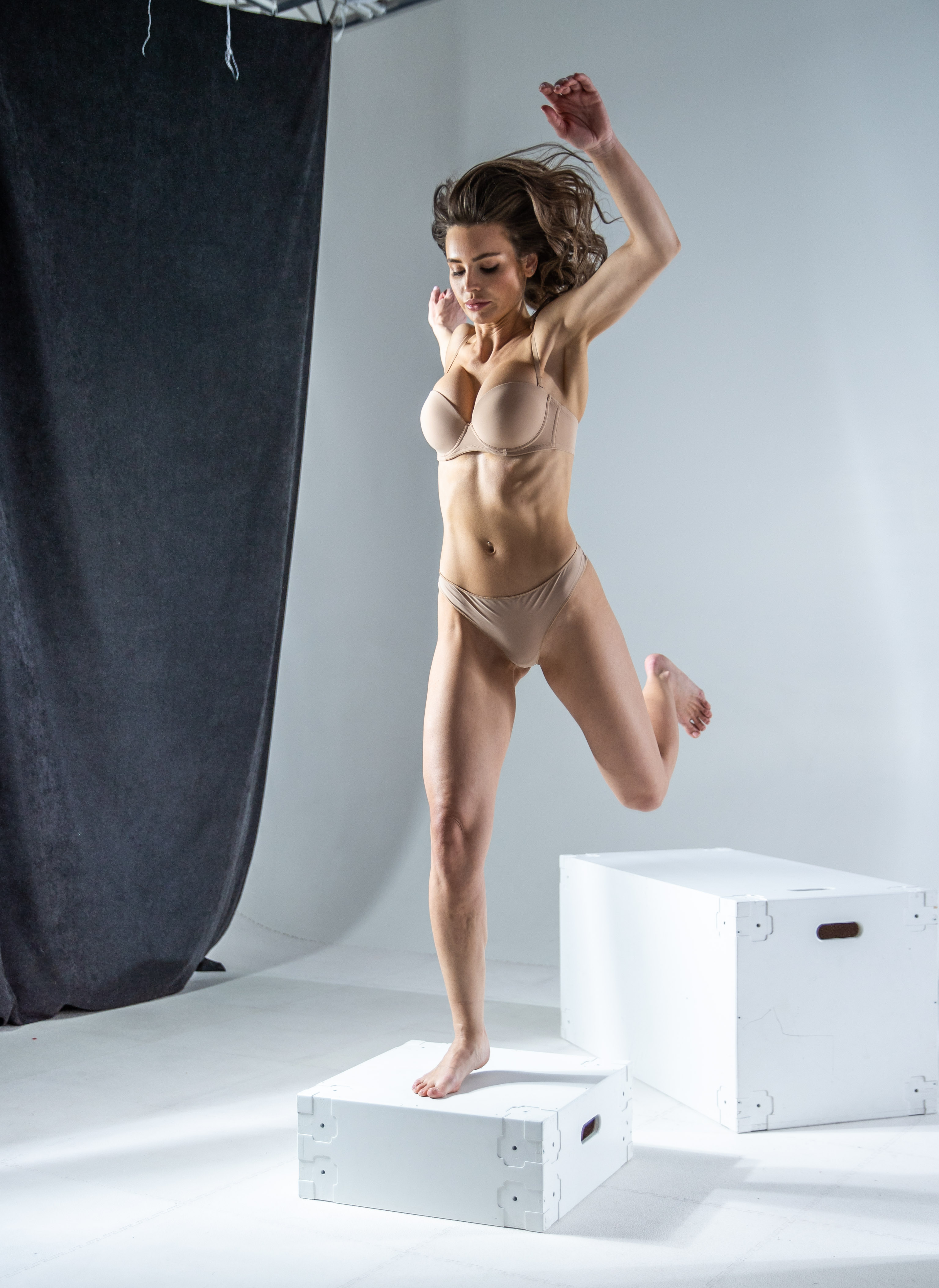 a woman in beige underwear jumps off a white box in a studio setting. there is another white box and a grey curtain in the background.