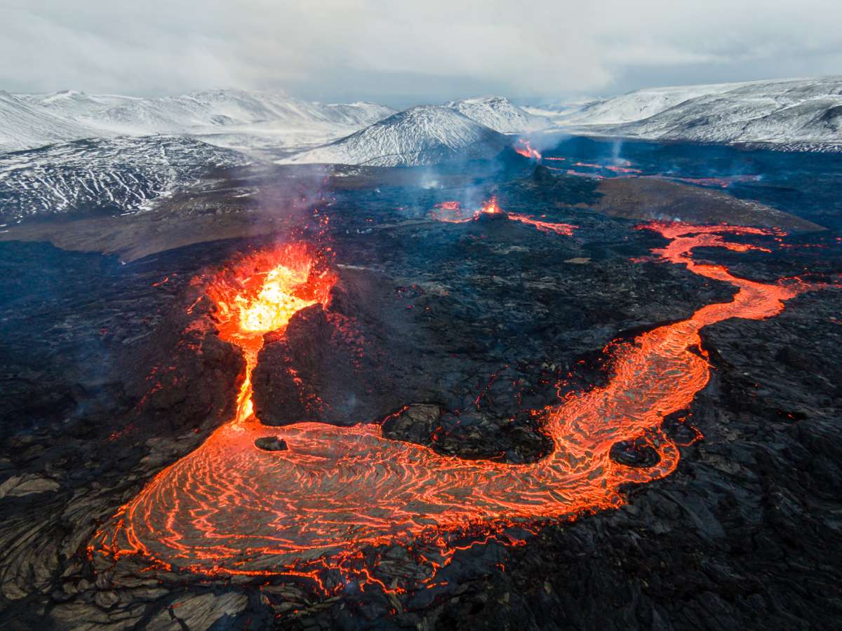 an aerial view of a volcanic eruption in iceland. molten lava flows from the crater of the volcano, creating a fiery river that cuts through the surrounding landscape. the lava is a bright red and orange color, and it is surrounded by dark, volcanic rock. in the distance, snowcapped mountains can be seen.