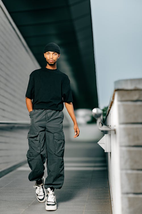 a stylish young man strolls confidently down a modern walkway. he's dressed in contemporary urban attire: a black beanie, a simple black tshirt, dark grey cargo pants, and white and black sneakers.  his posture and expression convey selfassurance. the setting is an outdoor urban environment, with clean lines of modern architecture forming the backdrop. the overall feel is one of effortless style and urban coolness.