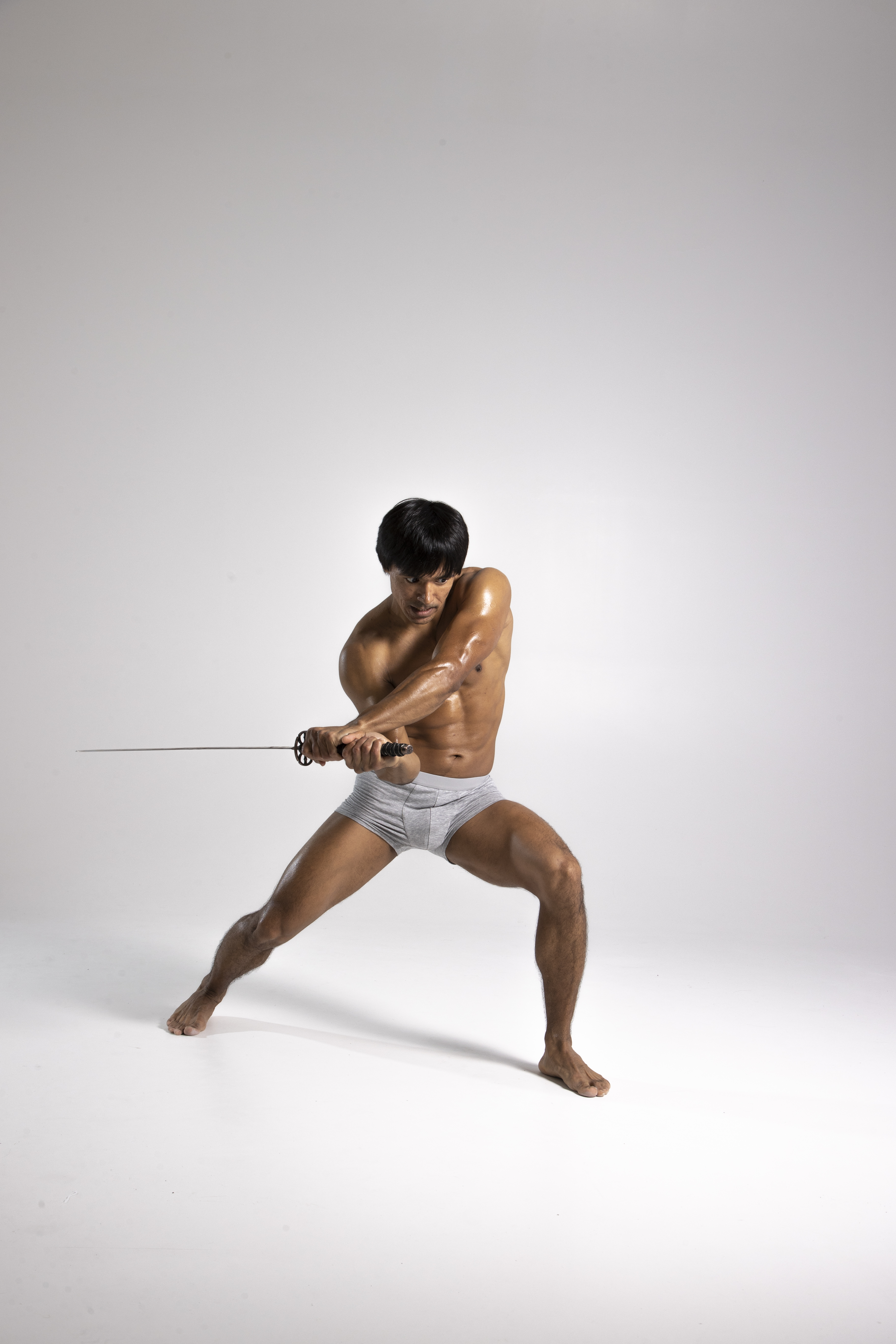 a muscular man in grey underwear is wielding a katana in a studio setting. he is in an action pose, demonstrating his physical strength and fitness. the sword is sharp and the man appears ready for combat or a martial arts demonstration.