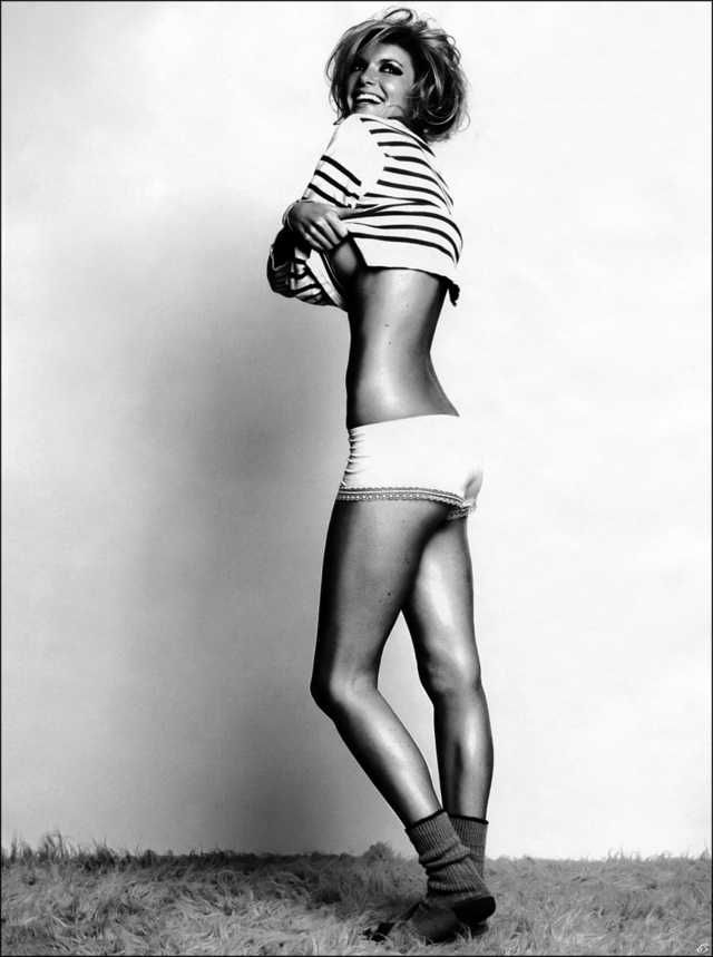 a black and white photograph of a woman standing in a studio setting. she is wearing white underwear and a striped top that she is pulling up, revealing her midriff. the lighting is bright and highlights her figure. the background is simple and white, drawing attention to the model and her pose.