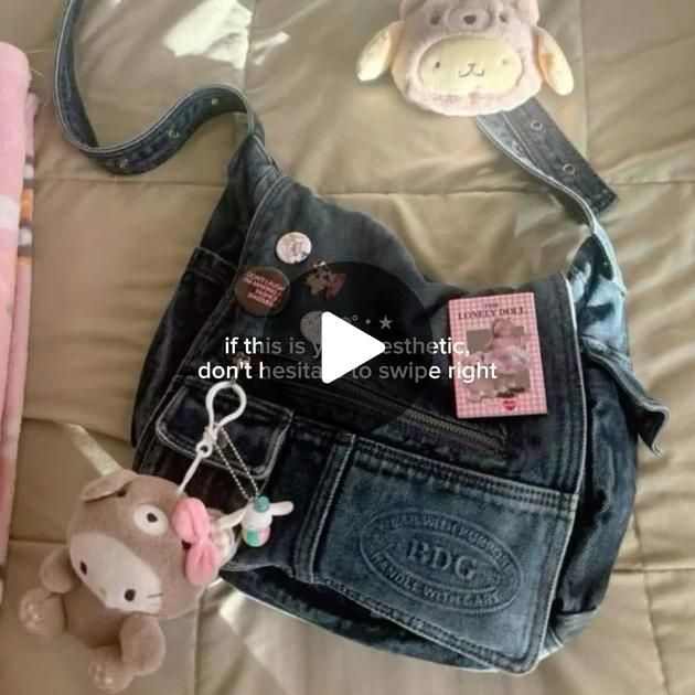 the image shows a denim purse decorated with various items. there's a small plush bear on top, and a brown doglike plushie hanging from a keychain on the left side of the purse. the purse itself has several buttons attached, including one with text. a "lonely doll" item is placed on the purse. the lower pocket of the purse has the "bdg" logo and the words "handle with care". a play button and text overlay are partially visible in the center.