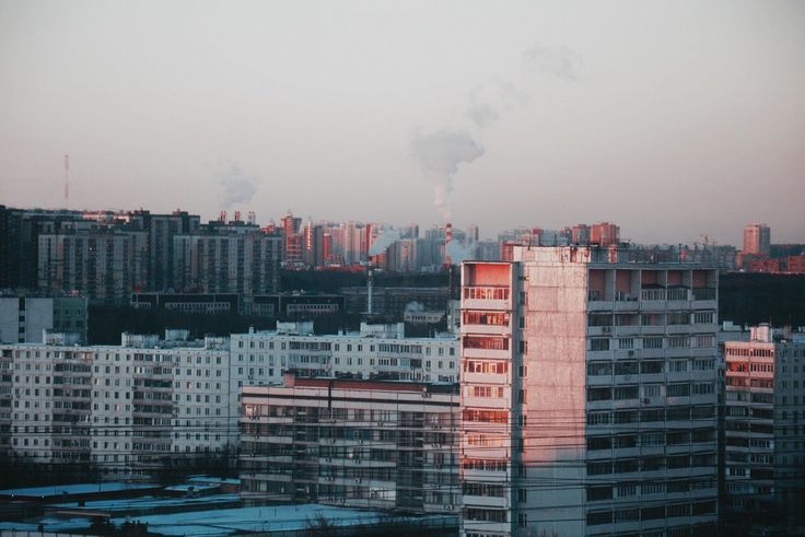 the image depicts a dense cityscape at either dusk or dawn.  numerous highrise apartment buildings, characteristic of sovietera architecture, dominate the scene. the buildings are predominantly light gray and white, with one taller structure displaying a pinkish hue from the setting/rising sun.  smoke plumes rise from industrial sources in the middle ground, adding an element of industrial activity to the image. the sky is a pale, muted color. the overall feel is one of a large, somewhat bleak yet peaceful urban landscape, possibly in a city in eastern europe or russia. there are hints of snow or ice on the ground in the foreground. power lines crisscross the view.