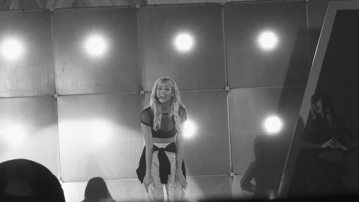 a black and white image shows a woman singing on a stage. she is wearing a midriffbaring top and pants, with a jacket tied around her waist. bright stage lights shine behind her. to her right, another figure stands in the shadows. the audience is visible in the foreground.