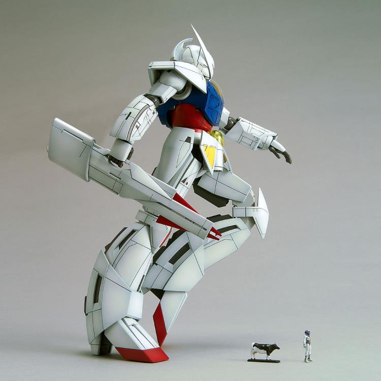a white, red, blue, and yellow gundam model kit stands tall in a dynamic pose. it's holding a large weapon in its right hand and has its left hand outstretched. a small figurine of a person stands to the right of the gundam, providing a sense of scale. a miniature cow also stands next to the human figurine. the background is a plain gray, and the lighting is soft.