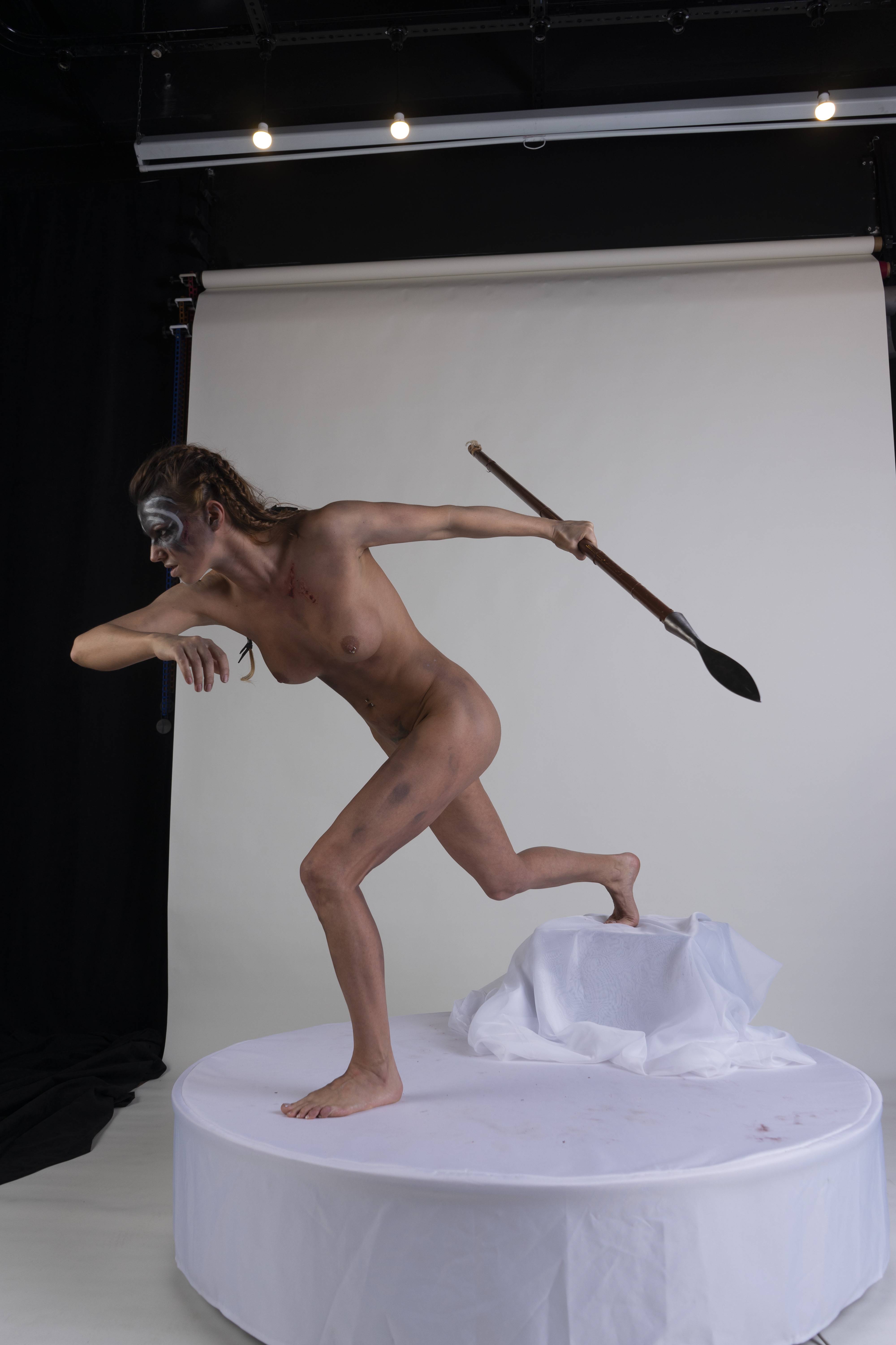 a nude woman with face paint and body paint in the style of a warrior is running with a spear in a studio setting. she is standing on a white platform in front of a white background.