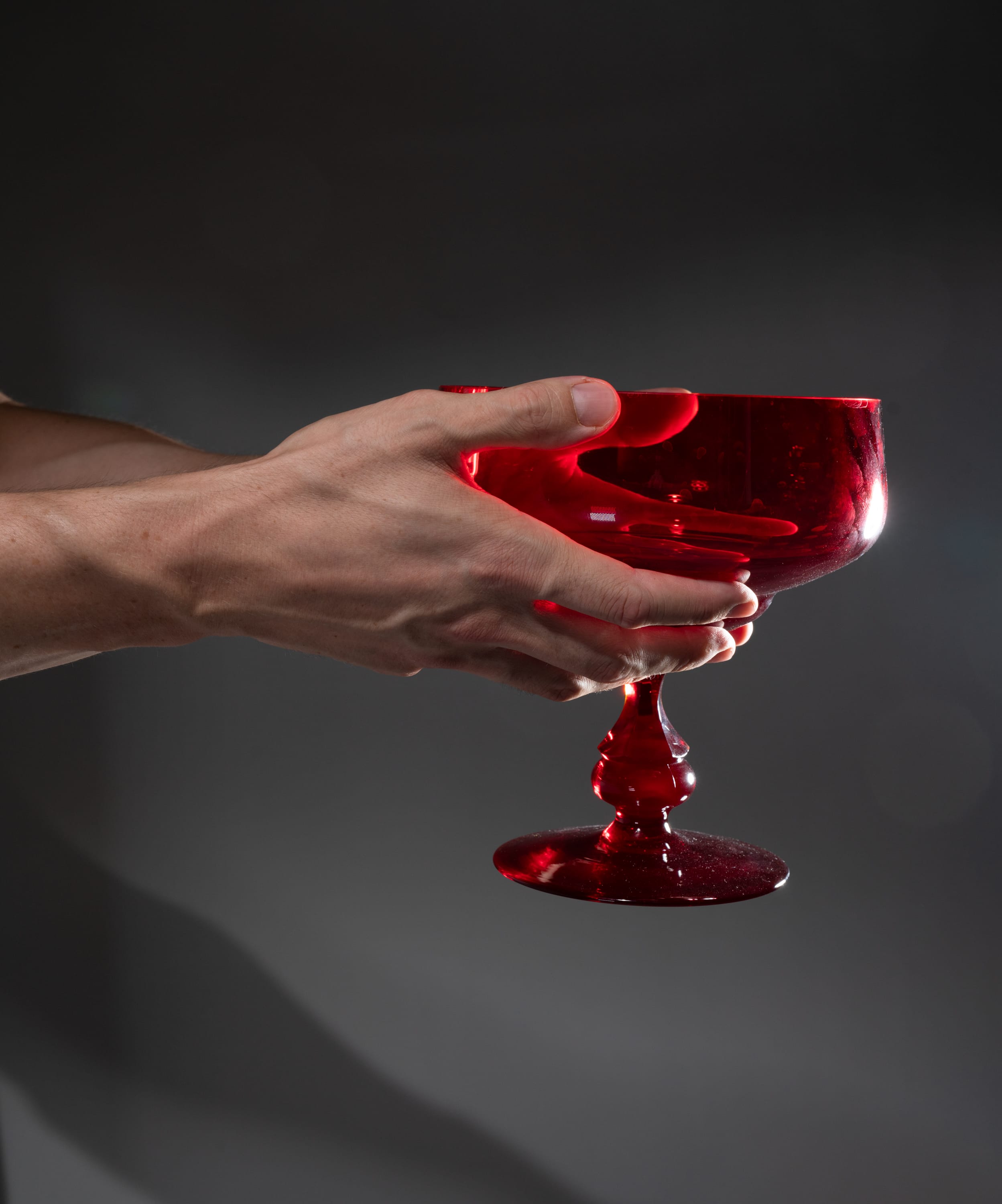 a man's hand gracefully holds a stemmed red glass against a gray background, illuminated by soft studio lighting. the image evokes a sense of elegance and sophistication.