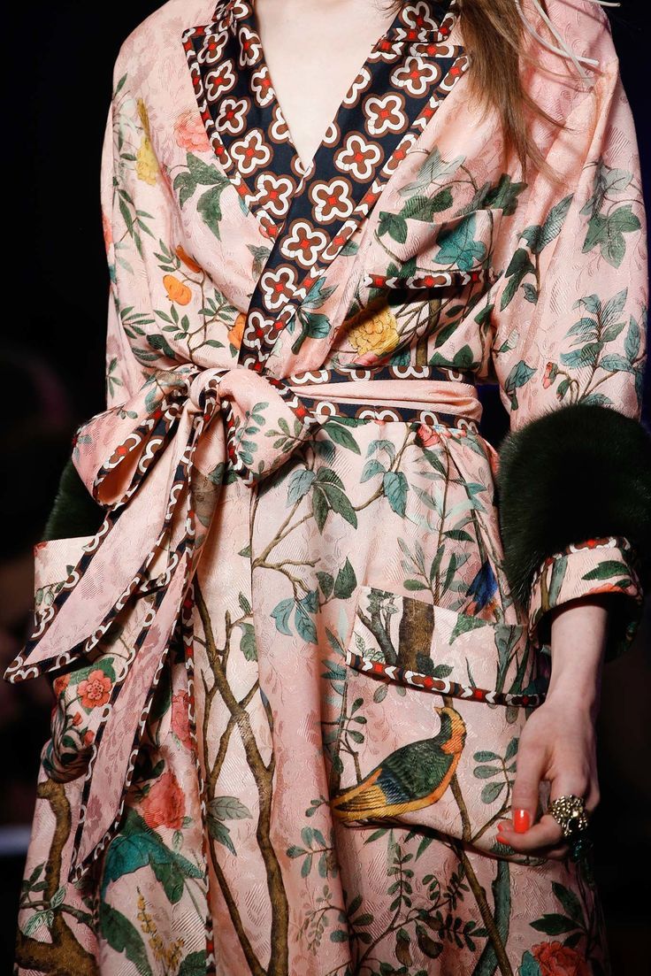 loseup shot of a model wearing a luxurious pink gucci robe. the robe features an elaborate floral print with birds and trees, detailed with a geometric pattern on the collar and belt. the sleeves are adorned with dark fur cuffs, and the overall style is elegant and opulent.  the image highlights the richness of the fabric and the intricate details of the design. the model's hand is visible, adorned with rings, showcasing the overall sophisticated aesthetic.