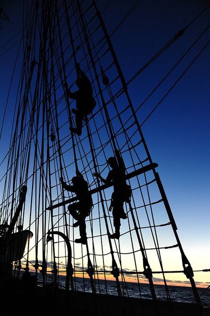 the image shows the silhouettes of three sailors climbing the rigging of a tall ship against a dark blue sky at dusk. the ropes and ladders of the mast are clearly visible, leading upwards. the ocean is seen at the bottom of the frame. the sailors are engaged in teamwork and adventure high up on the vessel.