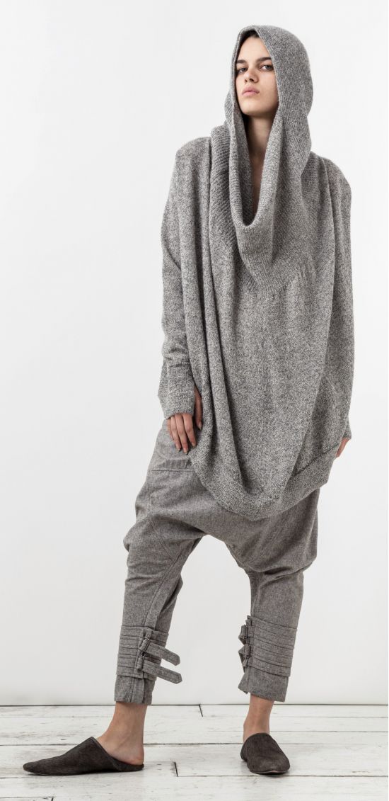 a full shot of a woman wearing a gray hoodie with a large cowl neck, gray harem pants with decorative straps at the ankles, and dark gray slippers, standing indoors in front of a white wall.