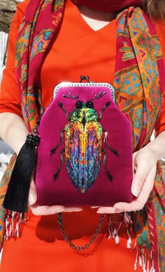 a woman in a vibrant red dress with a colorful scarf adorned with intricate patterns is holding a unique handbag, likely a clutch. the handbag is a bold statement piece, featuring a large, beaded and embroidered beetle design. the bag's vibrant colours complement the woman's outfit, showcasing a chic and elegant bohemian style. the overall image captures a sense of fashionforwardness and attention to detail.