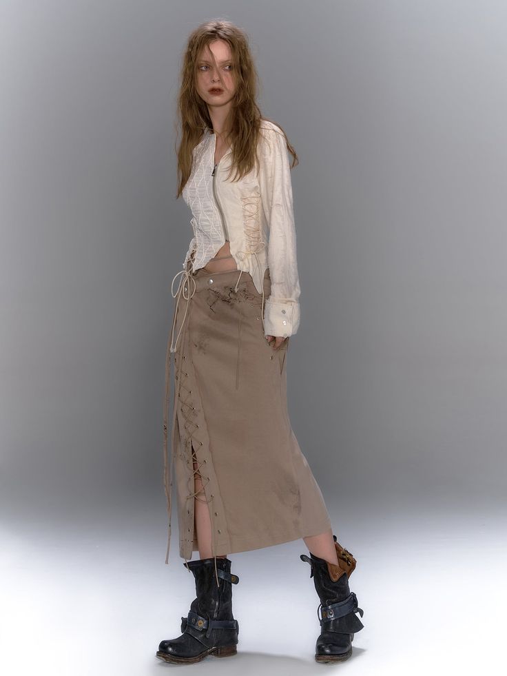 a fullbody studio shot of a female model posing in front of a light gray background. she is wearing an offwhite, distressed cropped top with laces and a long beige skirt with side lacing and slits.  she is styled with darkcolored, buckled ankle boots that have a grunge aesthetic.  the overall style of the clothing and pose conveys a bohemian or grunge fashion look.