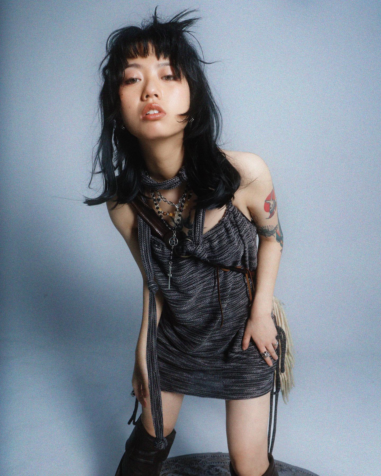 a full shot of a young woman with dark bobbed hair and bangs, wearing a short, dark grey knitted mini dress with long ties.  she's accessorized with layered necklaces and silver jewelry, and has visible tattoos on her arms.  she's also wearing high boots, partially visible. the model is posing with an intense yet fashionable gaze towards the camera, against a plain grey backdrop.  the overall style is edgy and alternative.
