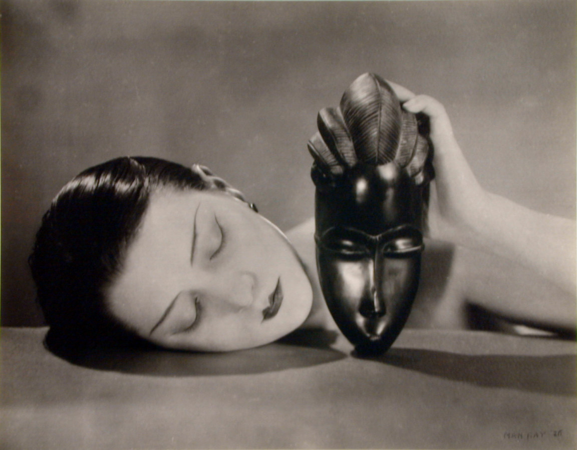 the black and white photograph depicts a woman with dark hair styled in a sleek, 1930s fashion, lying on her side with her eyes closed.  her head rests on a surface, and her arm is draped gracefully over a dark, carved african mask, which she seems to be gently holding or touching. the woman's expression is serene and peaceful, contrasting with the mask's powerful features. the lighting and composition are meticulously arranged, creating a mood that's both elegant and subtly unsettling, characteristic of surrealist photography. the photograph is a striking example of man ray's style, seamlessly blending portraiture and still life elements. the overall effect is one of dreamlike mystery and artistic sophistication.