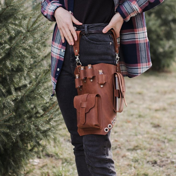 a person is wearing a brown leather belt bag on their right leg. the bag has compartments for vials and a larger pouch with a flap. it also has metal grommets along the bottom edge. the person is wearing dark gray jeans, a black shirt, and a plaid shirt over it. they have their hands on their waist. the background is blurry, but it appears to be outdoors with greenery.