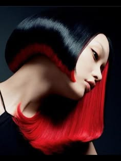 a woman with a red and black hairstyle looks over her shoulder at the camera. she has her hair styled in a bob and one side is dyed a bright red. her makeup is minimal and she looks straight at the camera. the photo is dark with a focus on the woman's hair.